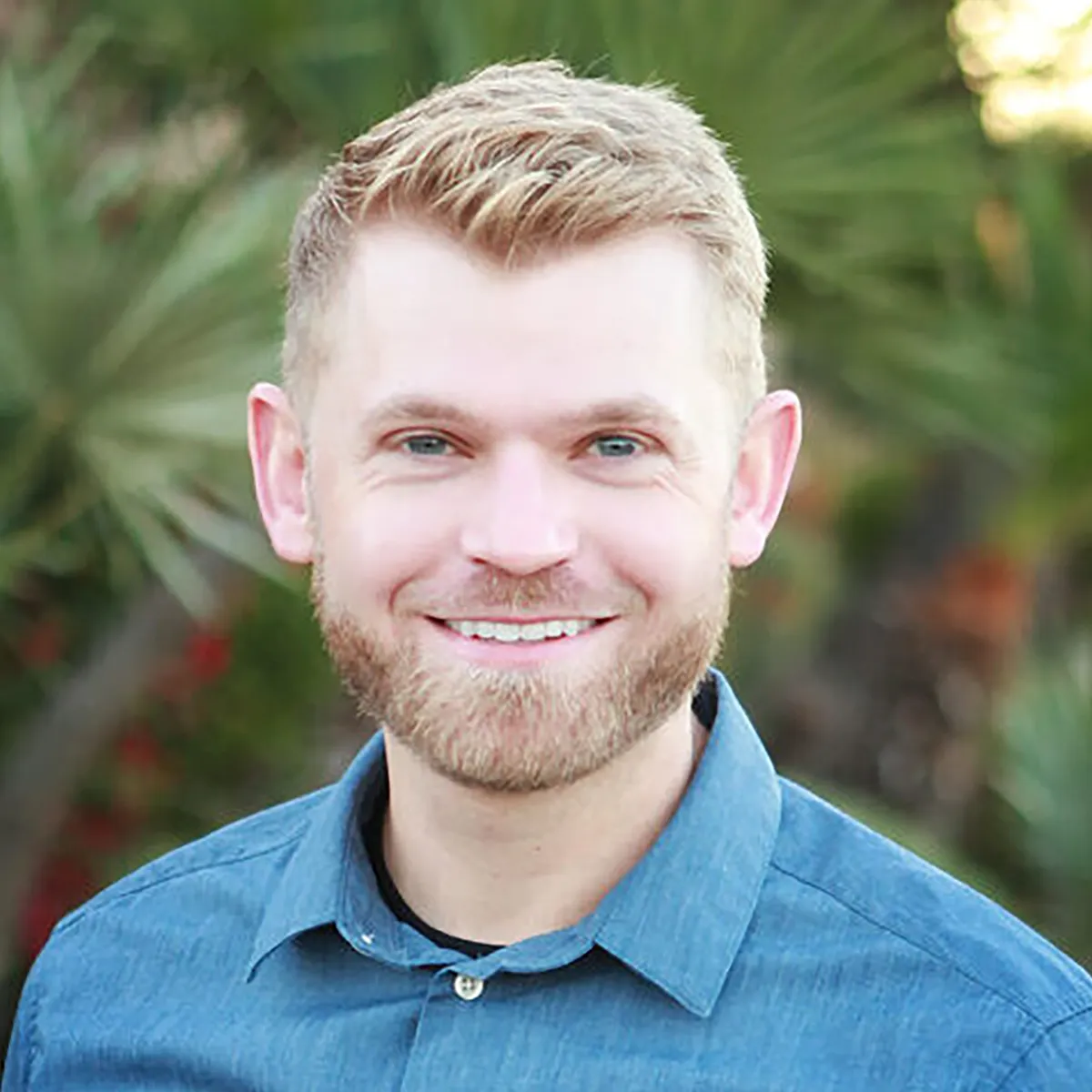 Professional headshot of smiling man in denim shirt against blurred green garden background.